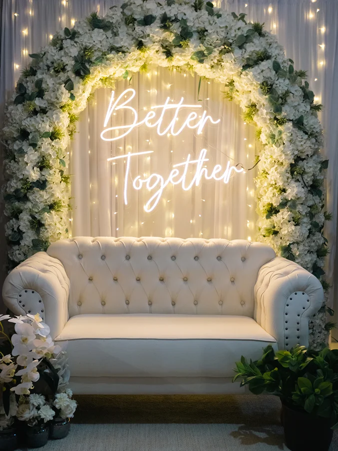 Foto ilustrando um espaço com um sofá de couro branco com um arco de flores no fundo do plano e uma placa de luz led. Representando a atenção do ASH Decorações para oferecer ambientes únicos para captar memórias, oferecendo uma vasta seleção de decorações para todos os gostos.