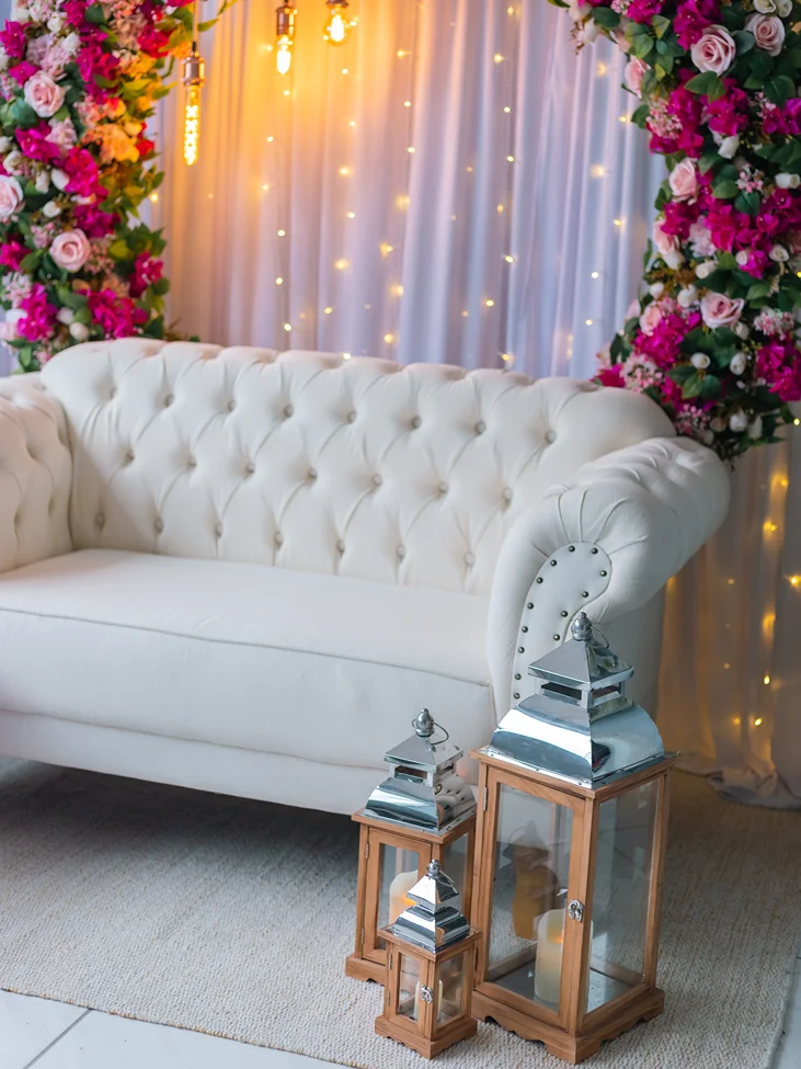 Foto ilustrando um espaço com um sofá de couro branco com um arco de flores no fundo do plano e uma placa de luz led. Representando a atenção do ASH Decorações para oferecer ambientes únicos para captar memórias, oferecendo uma vasta seleção de decorações para todos os gostos.